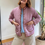 The Marley Edit pink quilted jacket with block print pattern, round neckline, button front, and tan binding. Model wears a blue sweater and beige trousers.