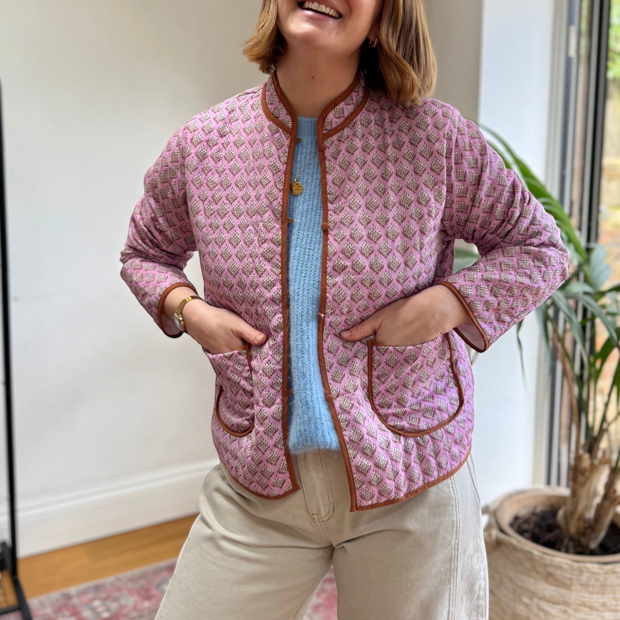 The Marley Edit pink quilted jacket with block print pattern, round neckline, button front, and tan binding. Model wears a blue sweater and beige trousers.