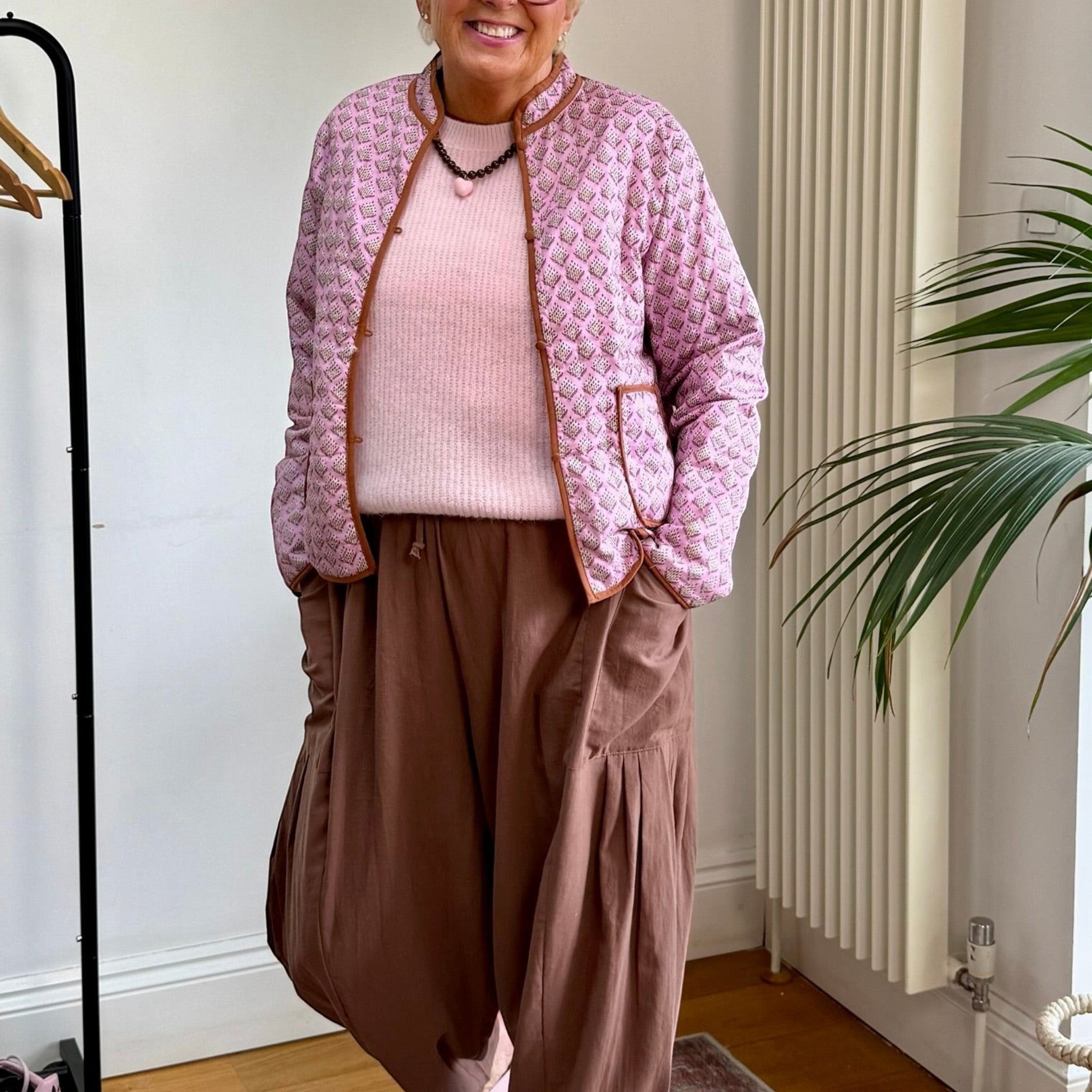 The Marley Edit pink quilted jacket with block print, tan binding, and button front, worn by a smiling woman.